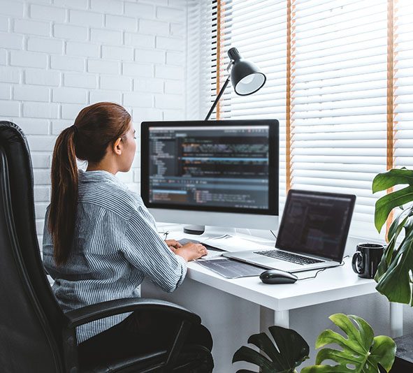 Person coding at a desk with a desktop monitor and laptop, surrounded by plants and natural light.